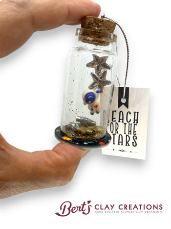 Hand holding a small glass bottle with stars and astronaut and a tag reading 'Reach for the Stars' by Bert's Clay Creations.